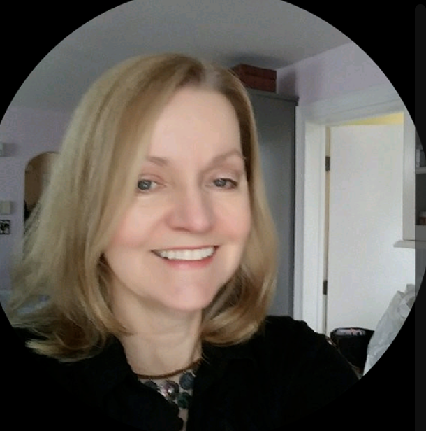 Selfie of a middle-aged woman who appears to be white with a warm smile wearing an elaborate necklace and a black dress shirt. She is inside the room of a well-lit house with white walls.