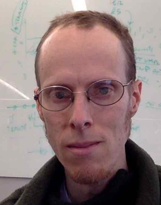 Photograph of a middle-aged male who appears to be white with short red-brown hair that’s thinner at the crown, side burns, a goatee, and oval wire-rimmed glasses in front of a whiteboard covered in formulas written with green whiteboard marker.