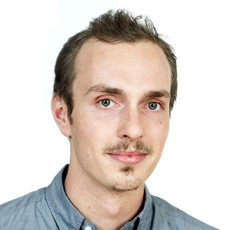 Headshot of a male who appears to be white with short brown hair, a light mustache and goatee. He has hazel-blue eyes and the top of his chambray blue button-up shirt is visible. The background is white.