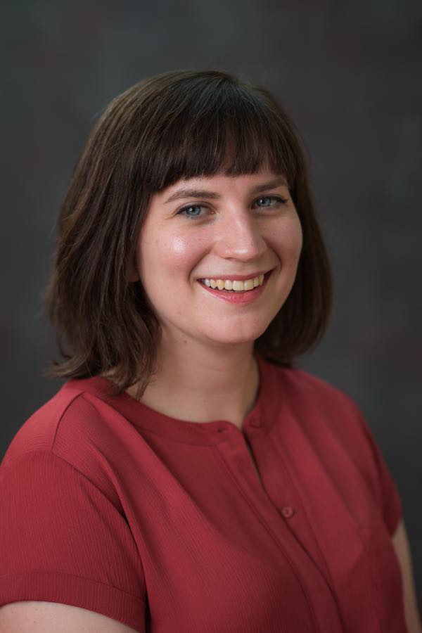 Portrait of a young woman who appears to be white with medium-length brown hair wearing a full smile and salmon-colored blouse in front of a gray backdrop.
