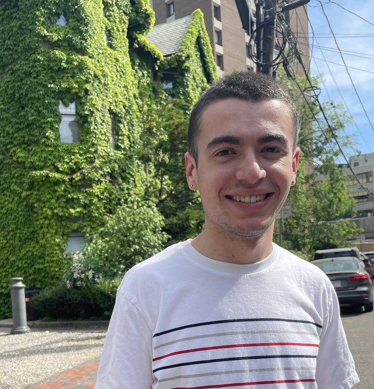 Photograph of a smiling young white man with short brown hair. He is wearing a white t-shirt with stripes, standing in front of a house covered in ivy on a sunny day.
