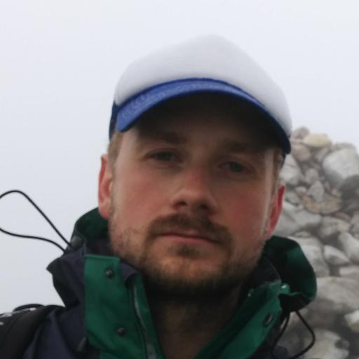 Close-up photo of a male who appears to be white  wearing a green and navy hooded outdoor jacket and a blue and white baseball cap. The background has white sky and rocks.