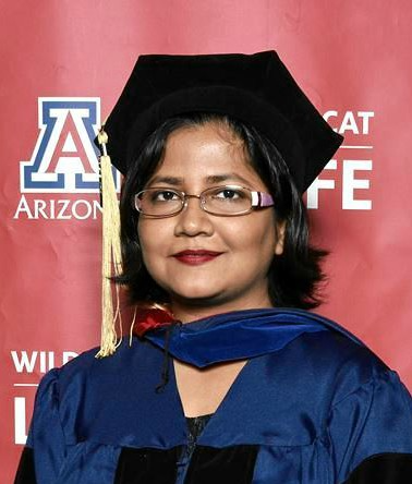 Photograph of a young woman who appears to be Asian with a warm smile wearing the doctorate regalia of the University of Arizona in front of a banner with the university’s colors and insignia.