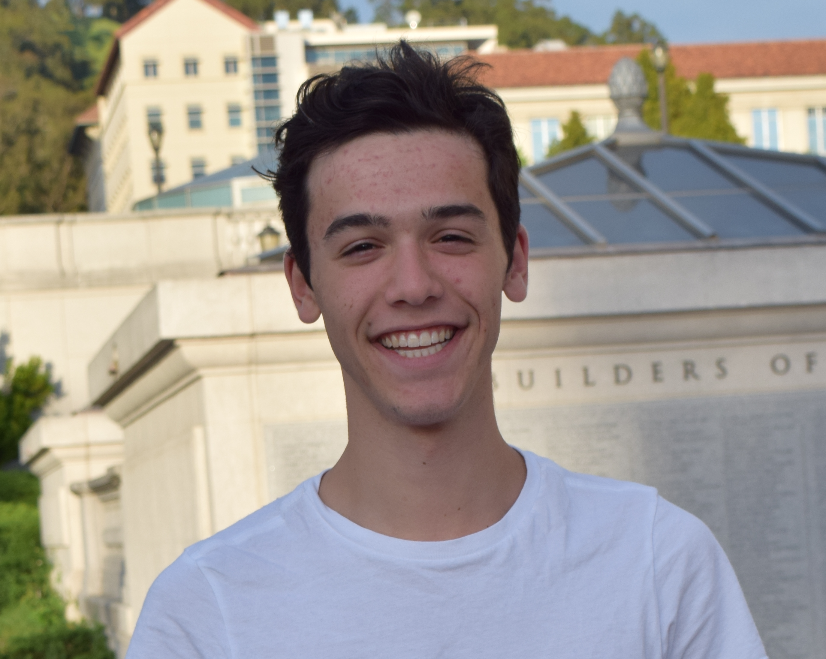 Headshot of a young male who appears to be white in front of a building. He’s got medium-short dark hair and is smiling broadly. He’s wearing a white t-shirt.