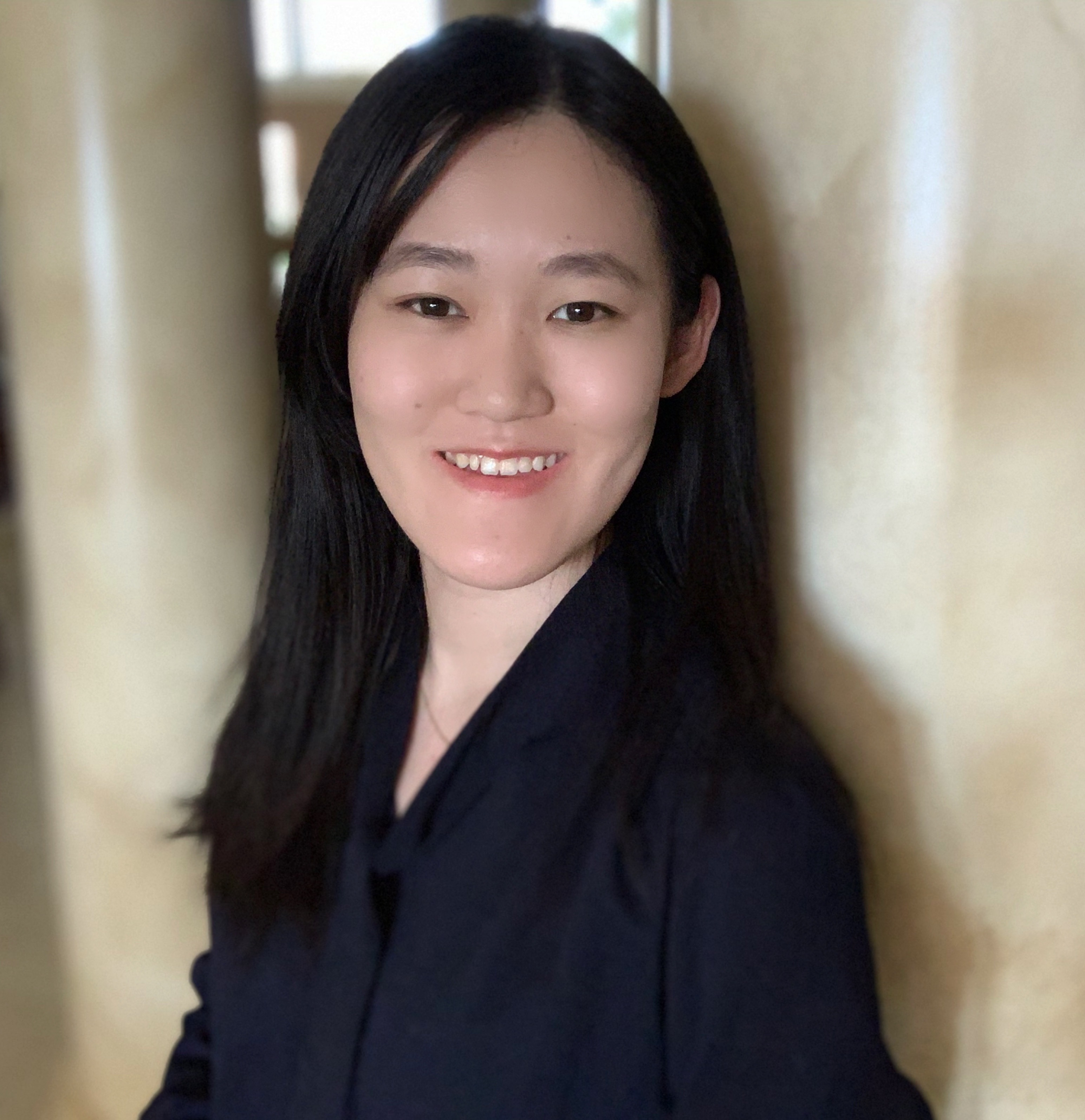 Headshot of a young Asian female with long, straight black hair and is smiling with an open smile. She is wearing a navy blue long-sleeve shirt, and is posing amongst marble columns inside a building.