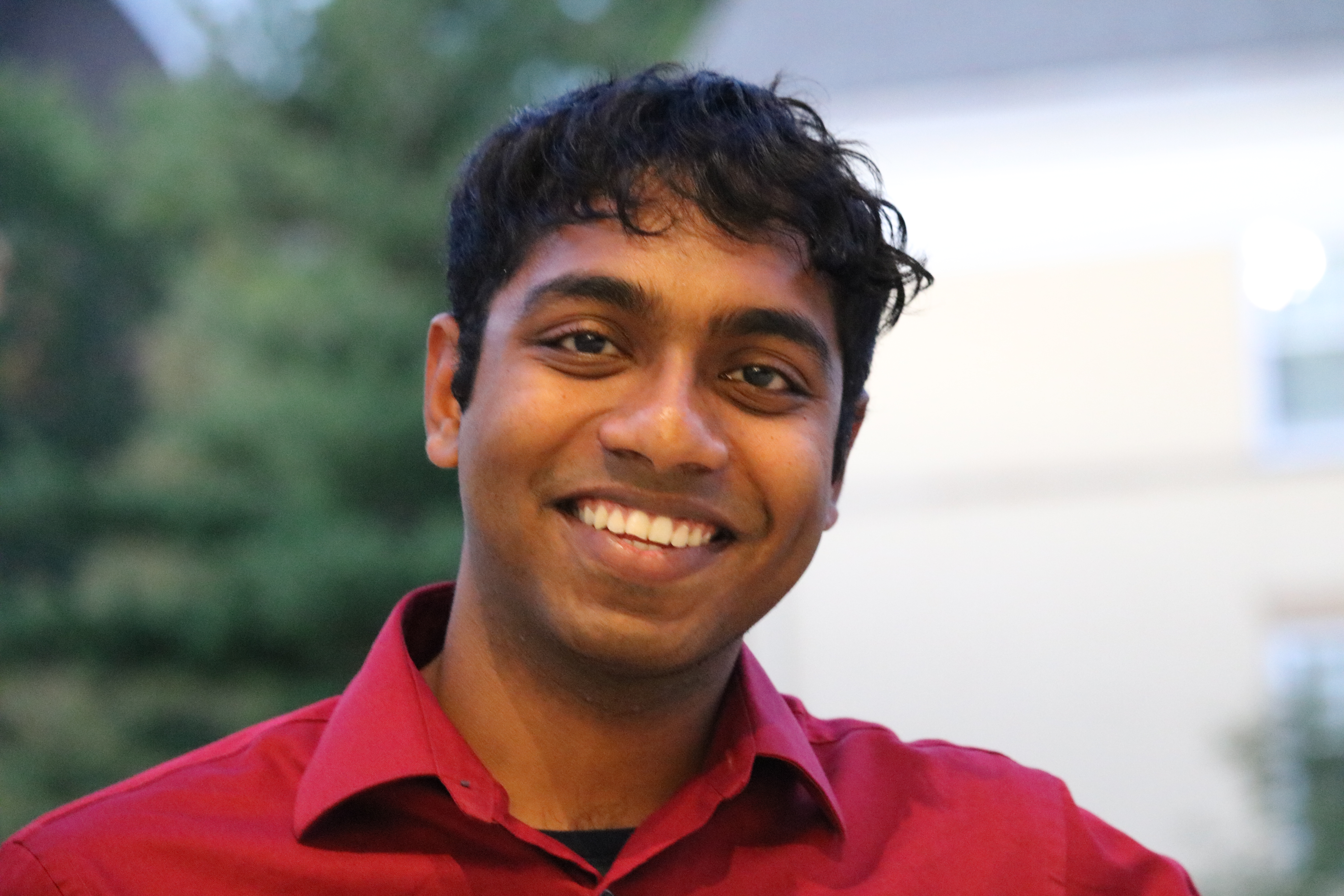 Photo of a smiling young man who appears to be Asian with wavy dark hair and brown eyes. He is wearing a red button-up collared shirt. The background is blurry with greenery and a white building.