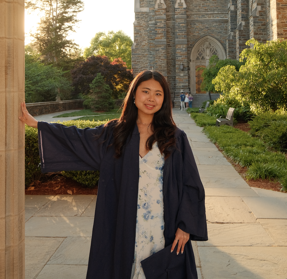 Portrait of a smiling young East Asian woman with long brunette hair.  She is wearing a dark blue gown over a floral dress, with gothic architecture and sunset in the background.