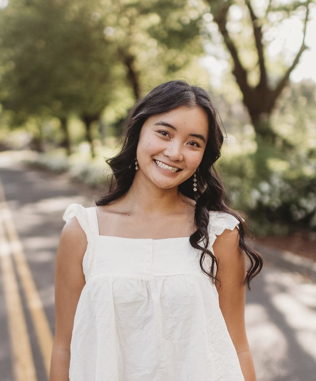 Portrait of a smiling young East Asian woman with long black hair.  She is wearing a white blouse, with trees and a road in the background.