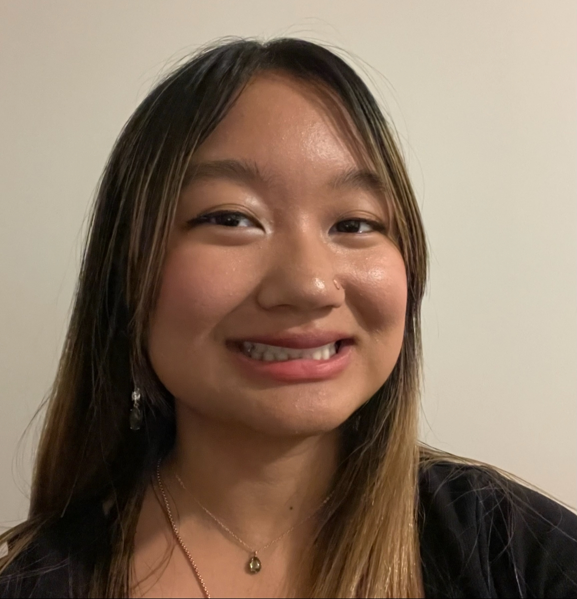 Headshot of a young woman against a neutral beige background. She appears to be smiling and has black hair with blonde highlights. She is wearing a black top, golden earrings and a necklace.