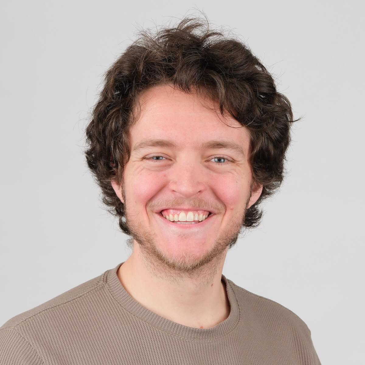 Headshot of a white man with medium curly brown hair and a short somewhat scruffy beard. He is smiling cheerfully in front of a neutral grey background.