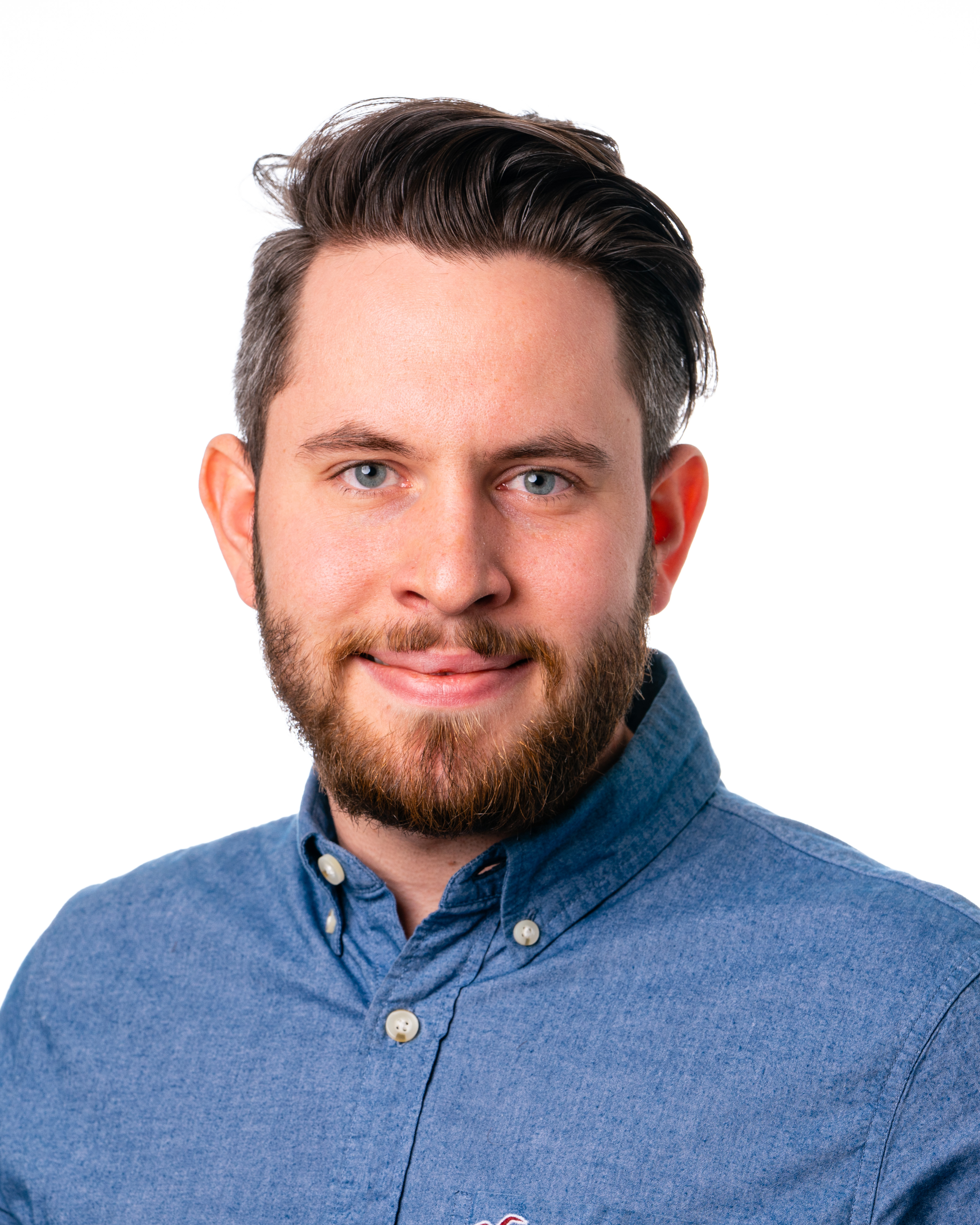 Headshot of a white man with medium long brown hair with hints of gray and a reddish-brown beard, wearing a blue shirt. The background of the image is white.