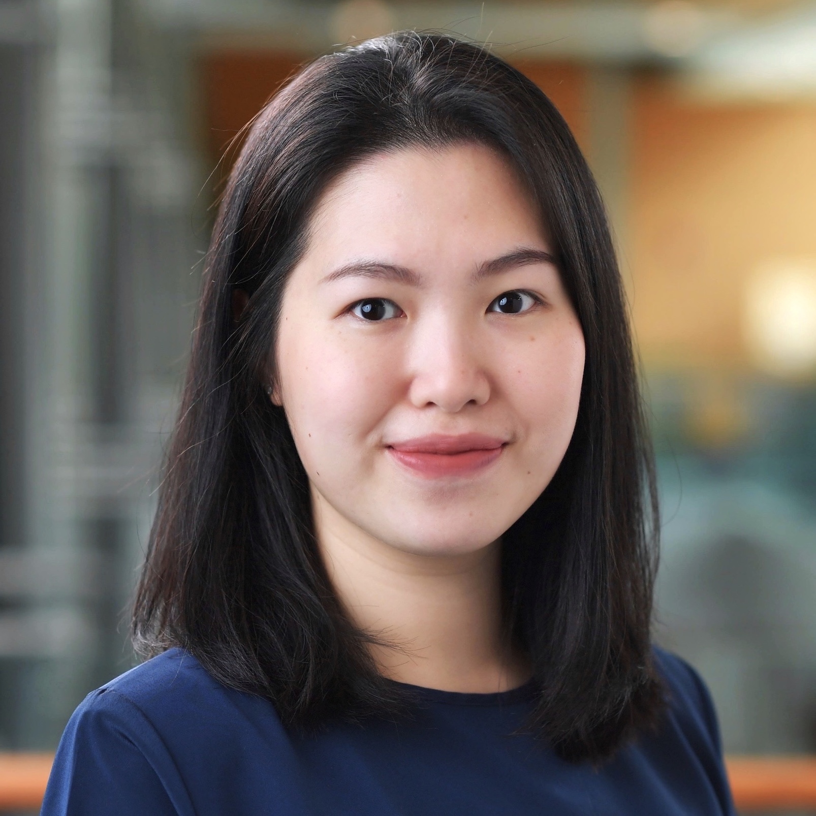 Headshot of a young Asian female smiling, with medium-length straight black hair, wearing a navy shirt in front of a colorful indoor blurred background.