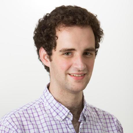 Photo of a young person with medium-length curly brown hair who appears to be white smiling in front of a white background. The person is wearing a plaid dress shirt and has curly brown hair.