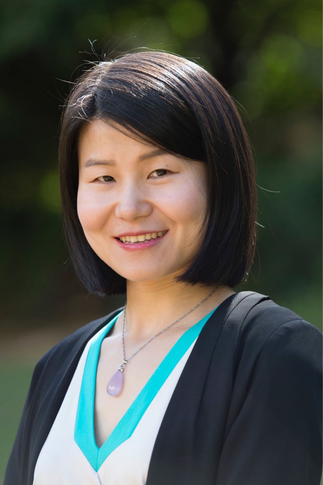 Photograph of a woman who appears to be Asian with medium-length straight black hair lightly smiling. The woman is outside in front of a blurred dark green background wearing a black academic gown over a white and teal top with a gemstone necklace.