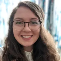 Headshot of a young white woman with a full smile and long, curly brown hair. She is wearing round browline glasses and a white button-up, standing in front of a blue glass background.