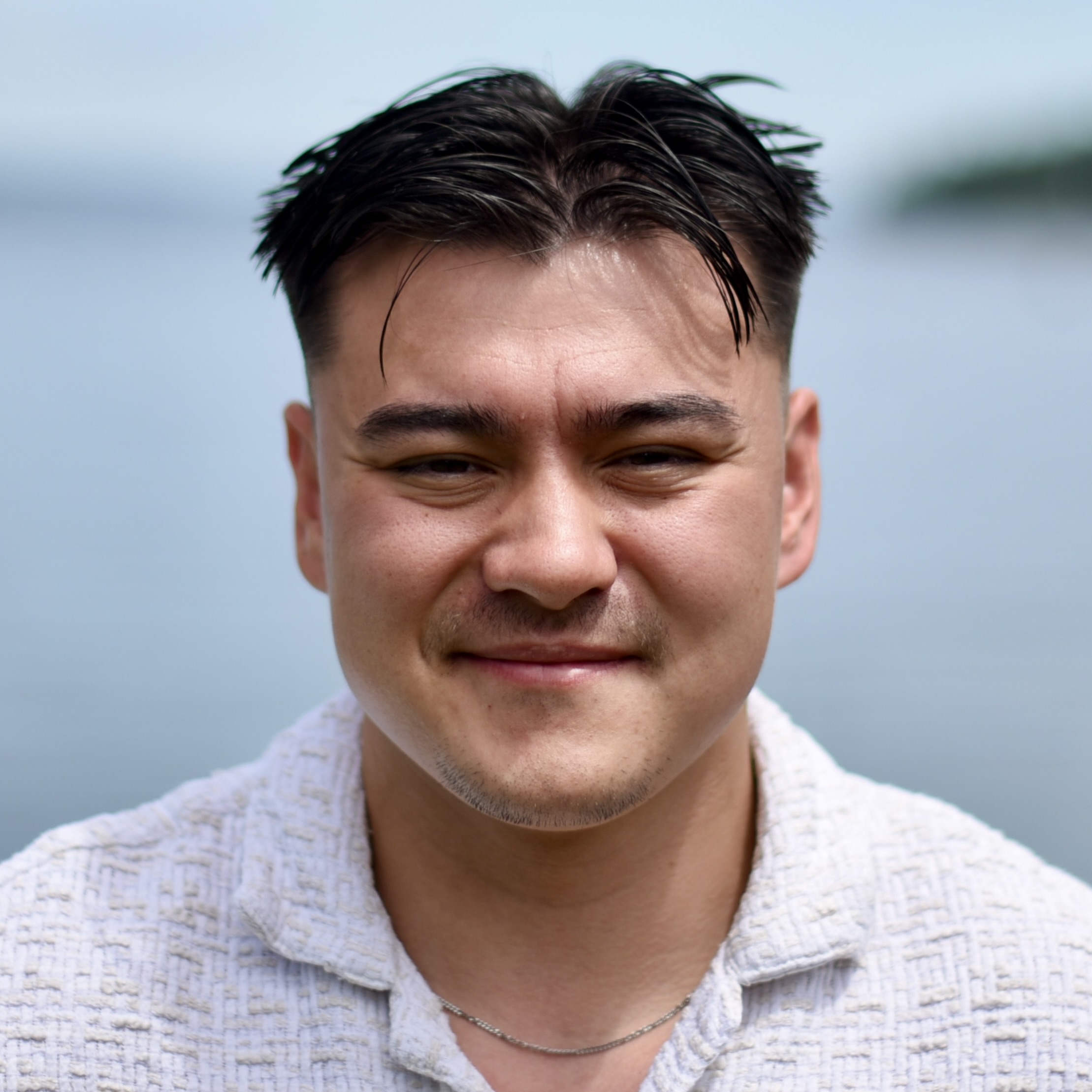 Photograph of a smiling young white man with short black hair. He is wearing a white collared shirt, standing in front of the ocean on a sunny day.