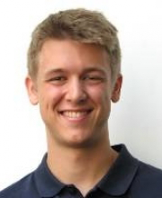 Photo of a young male who appears to be white with short blonde hair, smiling in front of a white background and wearing a navy polo shirt.