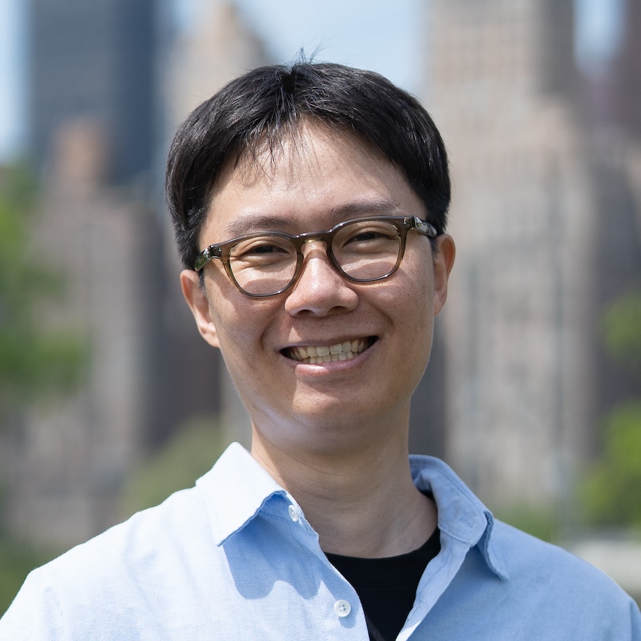 Photograph of an Asian man with short black hair and eyeglasses with a hint of a smile. He’s wearing a blue shirt in front of buildings and trees.