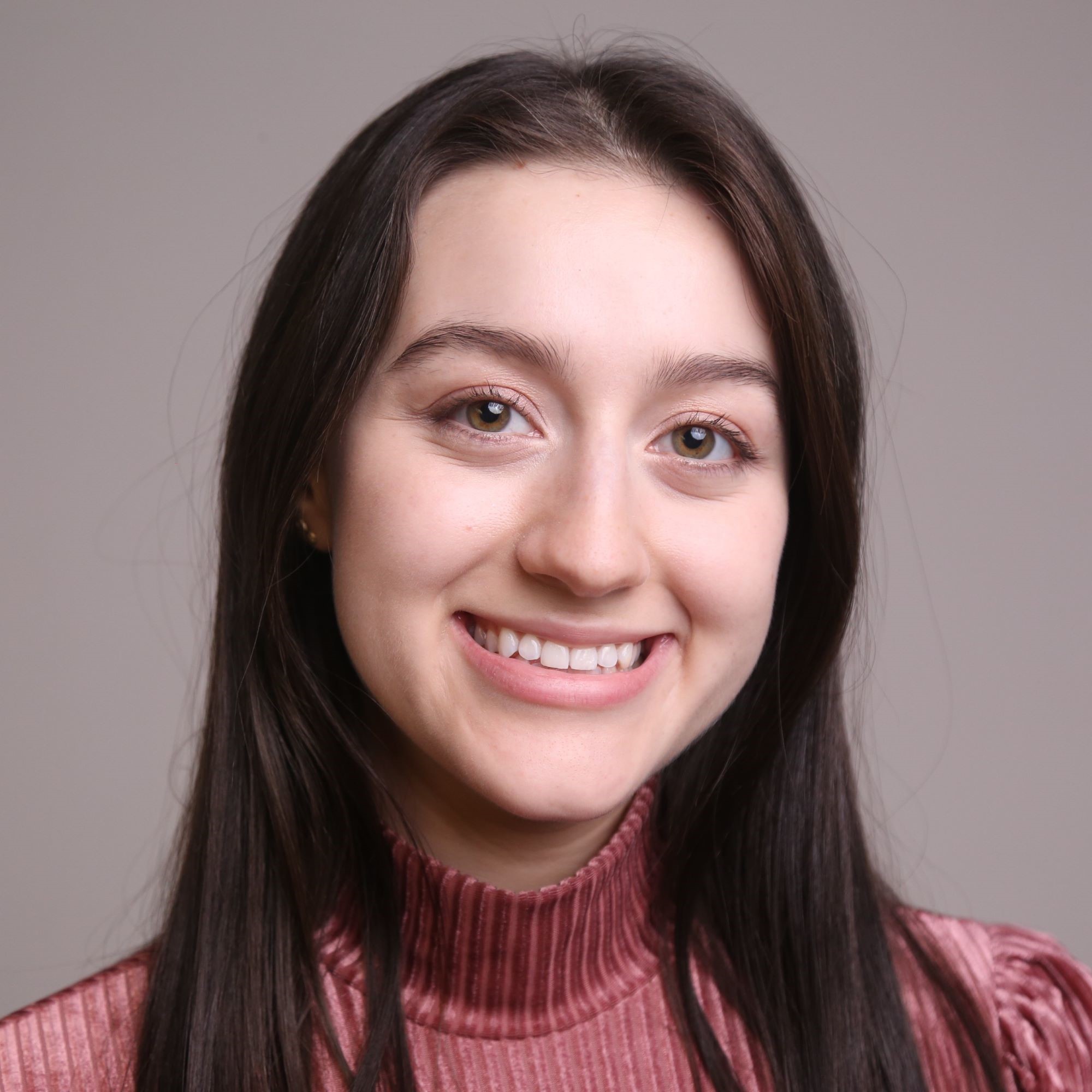 Headshot of a young white woman with shoulder-length brown hair and green eyes. She is smiling in front of a muted mauve background wearing a pink ribbed turtleneck shirt.