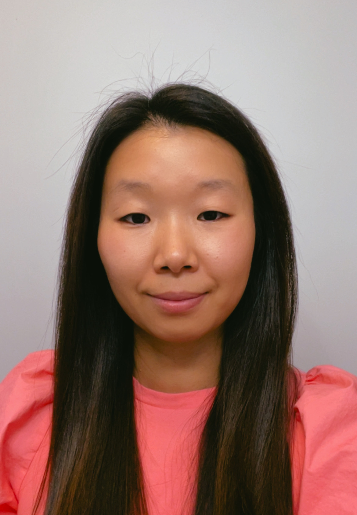 Photograph of young asian woman with long, straight black hair. She is wearing a bright pink top with a neutral expression. The background is a plain, light-colored wall.